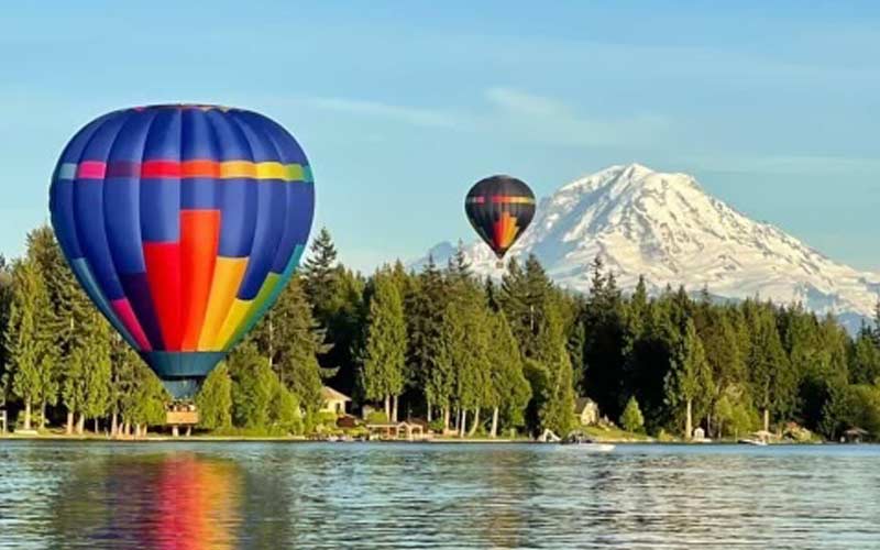 Two hot air balloons gracefully glide above a tranquil lake, framed by a majestic mountain
